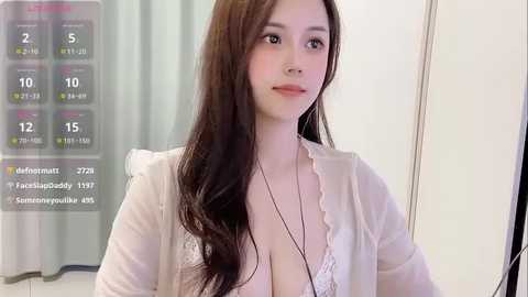 Media: Video of a young Asian woman with long brown hair, wearing a white lace bra, light-colored cardigan, and a medical bracelet, standing indoors, with a digital display showing vital signs in the background.