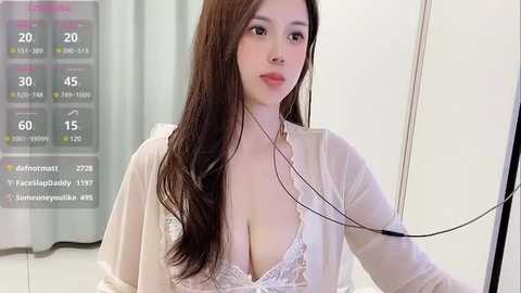 Media: Video of a young East Asian woman with long, dark hair, wearing a white lace bra, a sheer white robe, and headphones. Background shows a hospital room with a digital display and white walls.