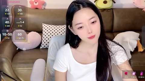 Media: Video of an East Asian woman with long black hair, fair skin, wearing a white t-shirt, sitting on a brown leather sofa, surrounded by plush toys and polka-dot pillows, with a digital camera display overlay.