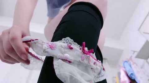 Media: Video of a person with fair skin wearing black leggings and a white lace-trimmed top. The individual is holding a pair of lace panties with pink floral patterns and bows. Background shows a blurred, brightly-lit room with white walls and a lamp.