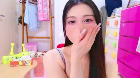 Video of an Asian woman with long black hair, wearing a pink tank top, holding a cigarette, in a cluttered, brightly lit room with toys and clothes.