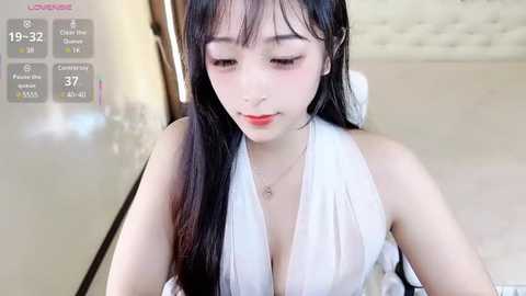 Media: Video of a young Asian woman with long black hair, wearing a revealing white halter top, in a modern, brightly-lit room with a tufted headboard.