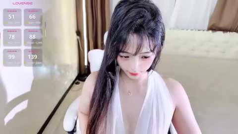 Media: Video of an Asian woman with long black hair, fair skin, and red lipstick, wearing a sleeveless white top, in a modern bedroom with a white bed and brown curtains.