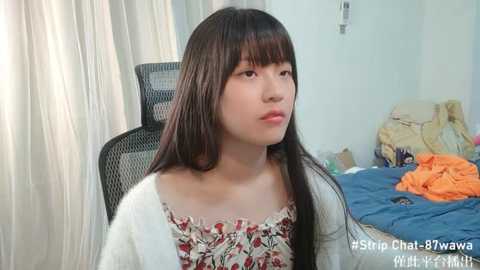 Media: Video of a young Asian woman with long black hair, wearing a floral-patterned top, sitting on a chair in a messy room with a blue blanket and clothes on the bed.