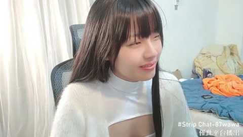 Media: Video of an Asian woman with long black hair, wearing a white turtleneck with a cutout revealing her cleavage, sitting on a black office chair in a messy bedroom.