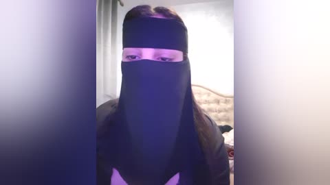 Media: Video of a woman wearing a black burqa, her face concealed, standing in a dimly lit bedroom with white walls and a bed.