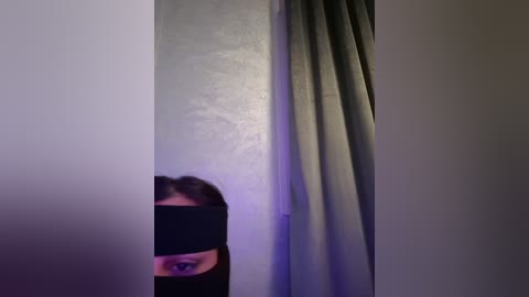 Media: Video of a person's face partially obscured by a black mask, with a gray curtain in the background, dim lighting.