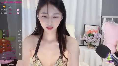 A video of an Asian woman with long black hair, fair skin, wearing a leopard-print bralette, standing in a bedroom with a floral arrangement, a bed, and a lamp.