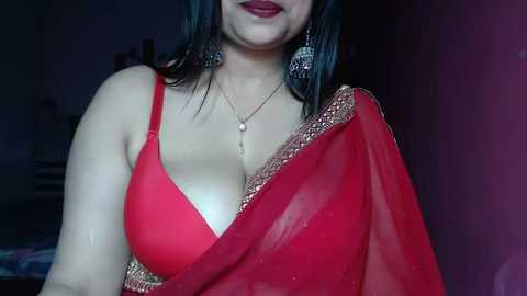 Media: Video of a woman with fair skin, wearing a red saree with a gold border, and a black blouse revealing cleavage. She has long, dark hair and wears large, silver earrings. The background is dimly lit with indistinct objects.