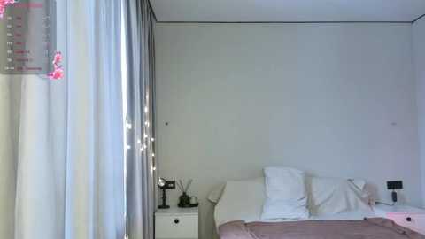 Media: A minimalist, modern bedroom with a white bed, white pillows, and a beige bedspread. On the left, a white nightstand with a black lamp and a pink floral decoration hangs on the wall.