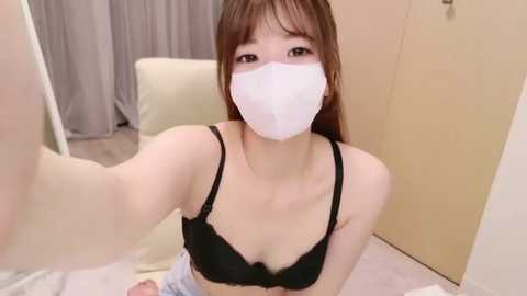 Media: Video of an Asian woman with long brown hair, wearing a black bra and white face mask, taking a selfie indoors against a beige background.