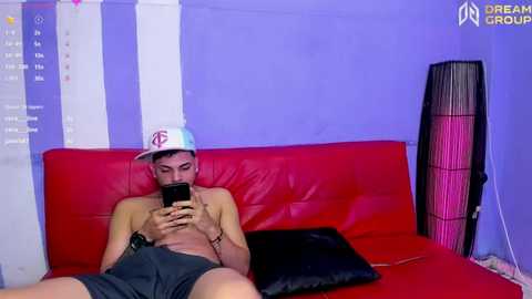 Media: Video of a shirtless, light-skinned man with short hair wearing a white hat and black shorts, sitting on a red couch, playing a smartphone game.