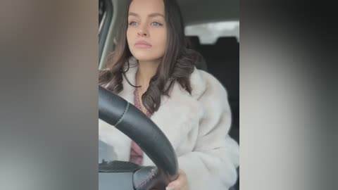 Media: Video of a young woman with long dark hair, fair skin, and light eyes, wearing a beige fur coat, driving a car.