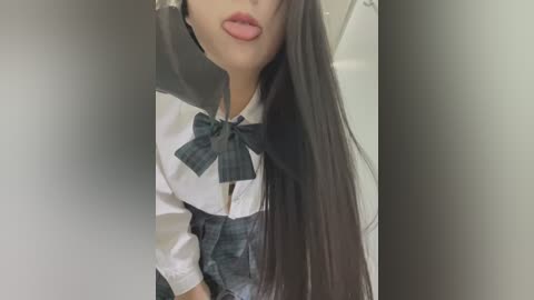 Media: Video of a young woman with long, straight, dark hair, wearing a white blouse with a large green plaid bow and a high-waisted pleated skirt. She is leaning forward, with her lips slightly puckered, creating a playful, close-up shot.