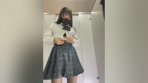 Media: Video of a young Asian woman in a schoolgirl outfit, consisting of a white blouse, black mask, and grey plaid skirt, standing in a narrow, dimly lit hallway with beige walls.