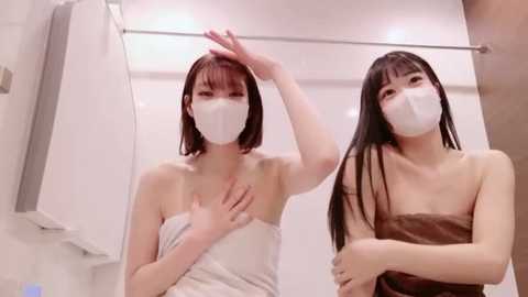 Media: Video of two Asian women with straight black hair, wearing white face masks, and wrapped in towels, standing in a modern bathroom with white tiles.