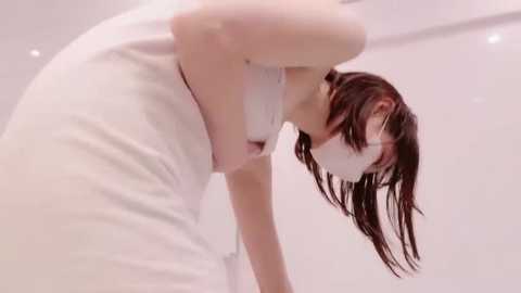 Video of a woman with long, wet brown hair, wearing a white dress, leaning over a bathtub. The background is a minimalist, white bathroom with soft lighting.