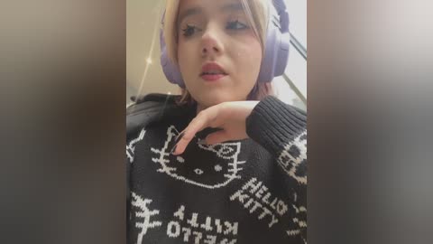 Media: Video of a young woman with light skin, blonde hair, wearing oversized purple headphones, black sweater with white text, and resting her chin on her hand.