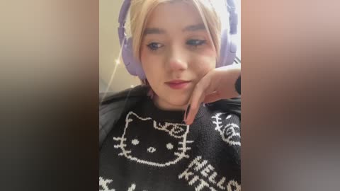 Media: Video of a young woman with light skin and blonde hair, wearing purple headphones and a black sweater with a Hello Kitty design, looking relaxed.