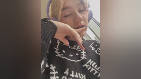 Video of a young woman with light skin, blonde hair, wearing a black sweater adorned with white cat motifs, resting her chin on her hand, eyes closed, with headphones on.