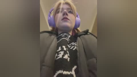 Media: Video of a young person with light skin and short blonde hair, wearing purple headphones, a black and white patterned scarf, and a puffy jacket, taken from a low angle, showing their face and upper body.