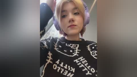 Media: Video of a young person with short blonde hair, wearing large purple headphones and a black sweater with white cat and \"Kitty\" text, standing indoors.