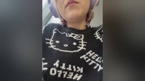 Media: Video of a woman wearing a black, patterned sweater featuring white Hello Kitty designs and Japanese text. She has fair skin, short pink hair, and is partially seen from the neck up.