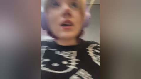 Media: A blurred video of a woman with blonde hair wearing a black t-shirt with a white skull design and large pink headphones.