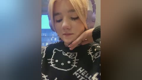 Media: Video of a young woman with light skin and blonde hair, wearing a black sweater with a white \"Hello Kitty\" design, resting her head on a large, fluffy, pink pillow.