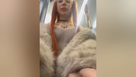 Media: Video of a young woman with orange pigtails, wearing a beige fur coat over a gray sweater, standing in a dimly lit subway car.