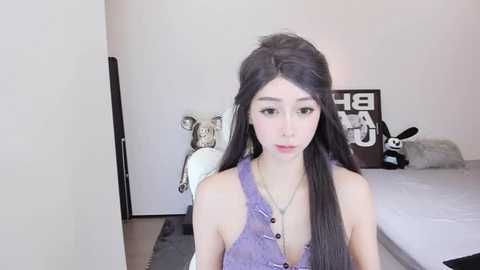 Media: Video of a pale-skinned, slender woman with long black hair in a ponytail, wearing a sleeveless purple top, standing in a minimalist bedroom with white walls, a bed, and a stuffed animal.