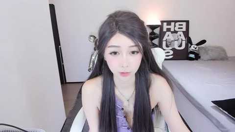 Media: Video of an East Asian woman with long, straight black hair, wearing a purple sleeveless top and silver necklace, sitting on a white chair in a modern, minimalistic bedroom.