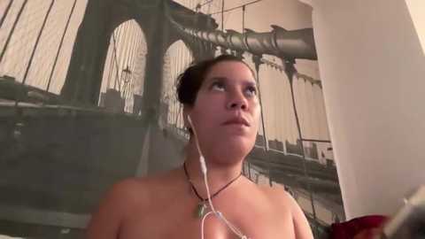 Media: Video of a topless woman with fair skin, dark hair in a bun, wearing white earphones, standing in a room with a mural of the Brooklyn Bridge.