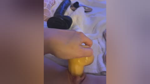 Media: A video of a close-up of a hand holding a yellow dildo against a person's vulva. The background features a bed with a white towel and a black vibrator.