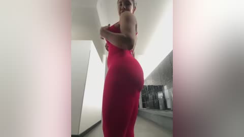 Media: Video of a woman in a red, tight-fitting dress, viewed from a low angle, showcasing her curvy figure. She stands in a modern, minimalist corridor with white walls and a metallic floor.