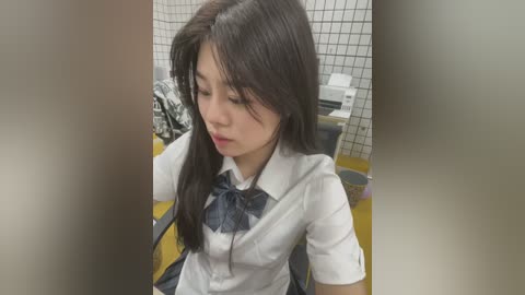 Media: Video of a young East Asian woman with long black hair, wearing a white blouse with a blue bow, seated in a public restroom with tiled walls and a yellow floor.