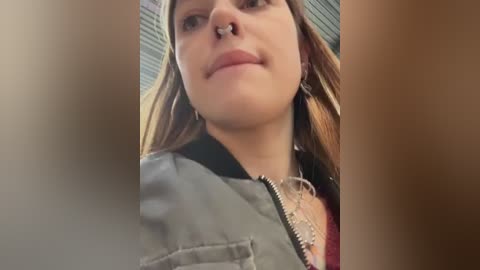 Video of a young Caucasian woman with fair skin and light brown hair, wearing a grey jacket and a silver nose ring, standing indoors with a blurred background.