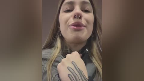 Media: Video of a young woman with light skin, long brown hair, and a septum piercing, smiling with her hands clasped. She wears a gray jacket and has tattoos on her hands.