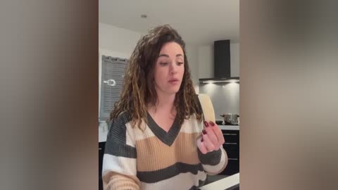 Video of a curly-haired woman in a striped sweater, holding a wooden spoon, in a modern kitchen with white walls and dark cabinets.