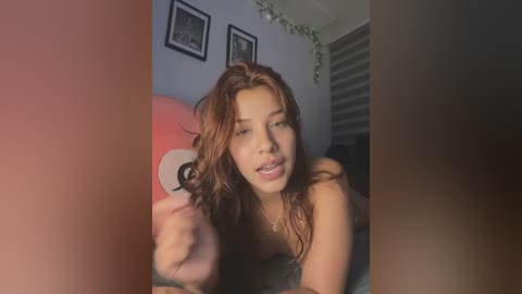 Media: Video of a topless young woman with wavy red hair, medium skin tone, and tattoos, smiling, lying on a bed with a gray blanket, surrounded by framed pictures and a string of lights.