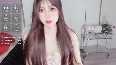 Video of an East Asian woman with long, straight brown hair, fair skin, and red lipstick, wearing a strapless pink dress, standing in a modern, minimalist room with a metal cart and white walls.