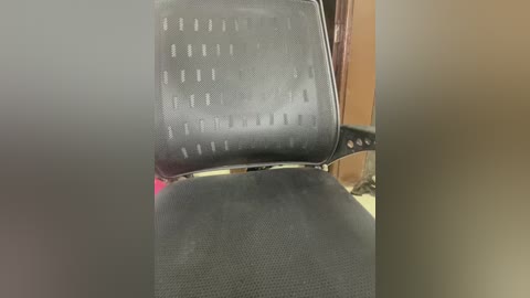 Media: A video shows a close-up of a black office chair with a textured fabric backrest and seat, slightly blurred, against a blurred background of a wooden desk and a wall.