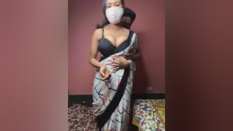 Media: Video of a South Asian woman in a black bra, white mask, and colorful sari, standing in a room with maroon walls, a bed with patterned bedding, and scattered items on the floor.