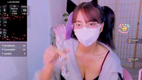 Media: Video of an East Asian woman with glasses, white mask, and black cat ears, wearing a light jacket, holding a clear plastic bag, in a dimly lit room with a gaming screen on the left.