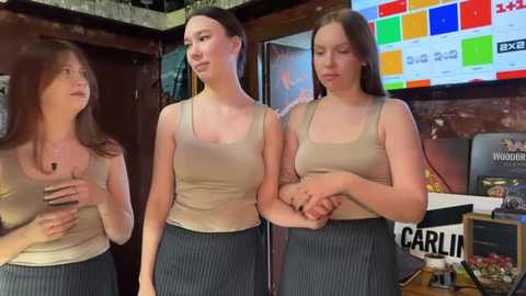 Media: Video of three young women in casual beige tank tops and gray pinstripe skirts, standing in a dimly lit bar with a colorful beer advertisement and neon lights in the background.