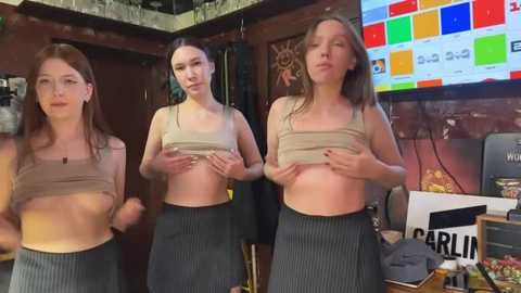 Media: Video of three young women with pale skin and long hair, wearing crop tops and pinstriped skirts, dancing in a dimly lit, cluttered room with a TV displaying colorful squares.