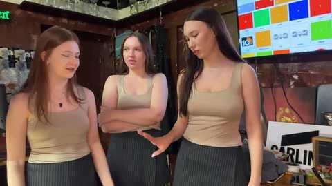 Video of three young women in casual beige tops and pinstriped skirts, standing in a modern bar with colorful wall graphics and glass shelves.