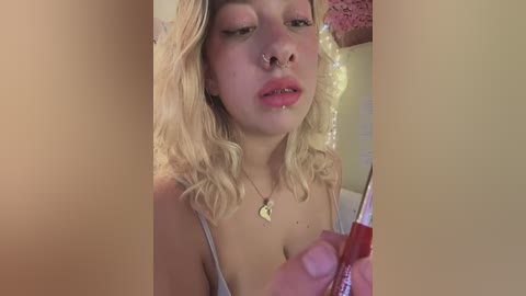 Media: Video of a blonde woman with a septum piercing, wearing a revealing white dress, applying lipstick in a dimly lit room with soft, pinkish lighting.