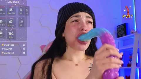 Media: Video of a young woman with long black hair and a black beanie, wearing a revealing top, licking a blue and pink dildo while in a bedroom with a white wall, a blue light, and a shelf with a TV.