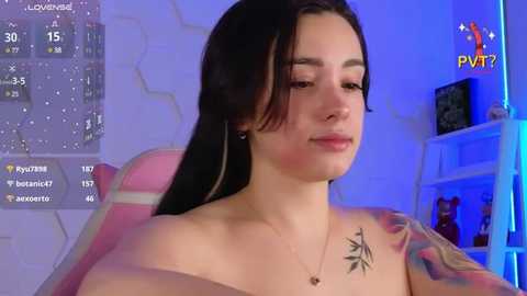 Media: Video of a topless woman with fair skin, dark hair, and a tattoo on her left shoulder, sitting in a pink gaming chair.